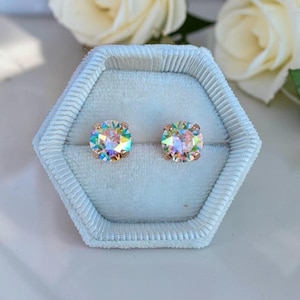 May include: A pair of round stud earrings featuring iridescent, multi-colored faceted stones set in rose gold metal. The earrings are displayed inside a light blue, hexagonal, velvet-lined jewelry box with a ribbed texture along the edges.
