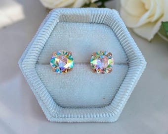 May include: A pair of round stud earrings featuring iridescent, multi-colored faceted stones set in rose gold metal. The earrings are displayed inside a light blue, hexagonal, velvet-lined jewelry box with a ribbed texture along the edges.