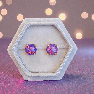 May include: A pair of round stud earrings with a gold setting. The earrings feature a vibrant, iridescent purple and pink opal stone. They are displayed in a white, hexagonal jewelry box with a soft, velvety texture.