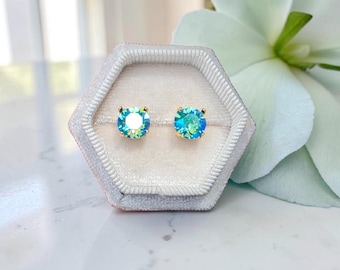 Può includere: Un paio di orecchini a lobo blu-verdi iridescenti con perni color oro, presentati in un astuccio esagonale di velluto grigio chiaro. Gli orecchini hanno un design rotondo e sfaccettato. Lo sfondo include un fiore bianco e una superficie di marmo.
