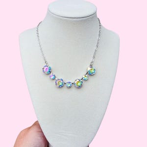 May include: A silver-toned necklace with iridescent, round gemstones of varying sizes is displayed on a white mannequin. The gemstones reflect a rainbow of colors. The necklace is on a pink background.