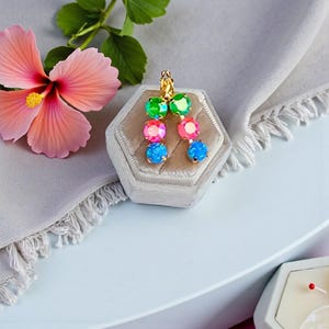 Puede incluir: Pendientes dorados con tres gemas redondas y coloridas en verde, rosa y azul. Los pendientes se exhiben en un joyero de terciopelo gris, con una flor de hibisco rosa y hojas verdes en el fondo.