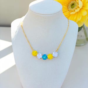 May include: A gold chain necklace with five colourful gemstones. The gemstones are yellow, white, and turquoise. The necklace is displayed on a white mannequin bust. A yellow flower is in the background.