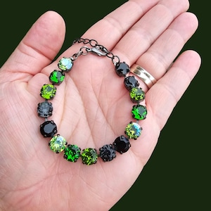 Pode incluir: Uma pulseira com corrente e fecho de metal escuro. A pulseira apresenta uma série de pedras preciosas redondas e facetadas em tons de verde, preto e um toque de azul. As pedras são espaçadas uniformemente ao longo da corrente.