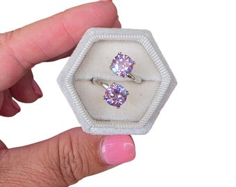 May include: A bypass ring featuring two round-cut, light purple stones set in silver-toned metal. The ring is displayed inside a hexagonal, light gray velvet ring box with a soft, plush interior lining designed to hold jewelry securely in place.