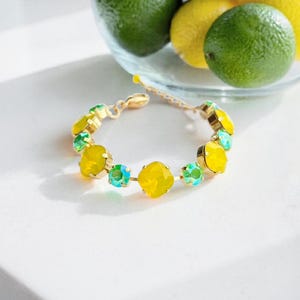 Op de afbeelding: Een goudkleurige armband met afwisselend gele en turquoise edelstenen. De armband is cirkelvormig en ligt op een wit oppervlak, met een glazen kom met limoenen en citroenen op de achtergrond.