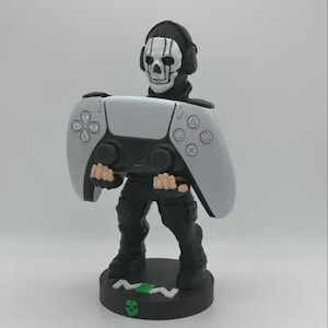 May include: A white and black figurine of a video game character holding a white PlayStation 5 controller. The character is wearing a black and white skull mask and has a black and white headset. The figurine is standing on a black base with a green logo.