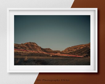 May include: A photograph of a desert landscape with a road in the foreground. The sky is a deep blue, and the mountains are a reddish-brown colour. The road is lined with red lights, which are likely from cars driving through the scene.