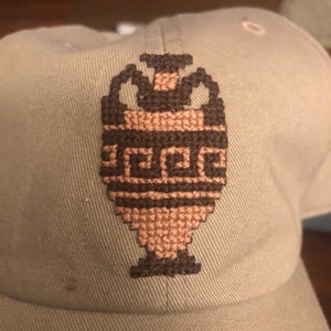 Könnte beinhalten: Eine graue Baseballkappe mit einem braunen und rosafarbenen gestickten Design einer antiken griechischen Urne.