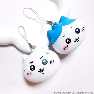 May include: Two cartoon character keychains. One keychain is white with a bear-like face, and the other is white with a blue cat-like face. Both have black eyes, pink cheeks, and a silver lanyard.