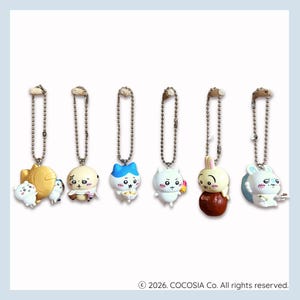 May include: Six small character keychains with silver ball chains. The figures include a white creature with a fish, a round beige character, a blue-eared white cat, a white creature with pink cheeks, a rabbit on a brown sphere, and a white rabbit.