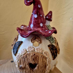 Könnte beinhalten: Eine Keramikfigur in Form eines Pilzhauses mit einem glänzenden roten Hut mit weißen Punkten. Der weiße Sockel hat eine strukturierte braune Bogentür mit herzförmigem Ausschnitt, steinartige Details um den Eingang und dunkelbraune Akzente.