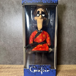 May include: A doll of a character from the movie "Coraline" is displayed in a clear plastic box. The doll wears a red jacket with orange polka dots, black pants, and glasses. The box has the movie title "Coraline" in white script.