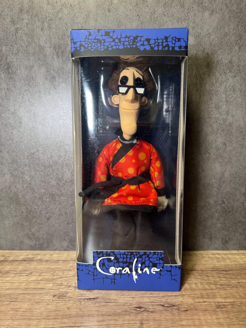 May include: A doll of a character from the movie "Coraline" is displayed in a clear plastic box. The doll wears a red jacket with orange polka dots, black pants, and glasses. The box has the movie title "Coraline" in white script.