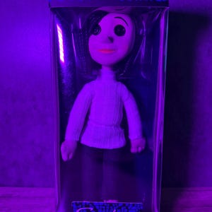 May include: A doll of a character from the movie "Coraline" in a clear plastic box. The doll has black hair, a white sweater, and dark pants. The box has the word "Coraline" printed on it.