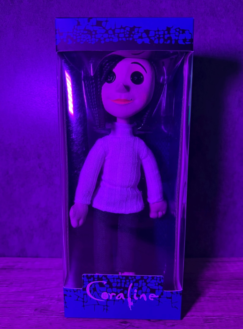 May include: A doll of a character from the movie "Coraline" in a clear plastic box. The doll has black hair, a white sweater, and dark pants. The box has the word "Coraline" printed on it.