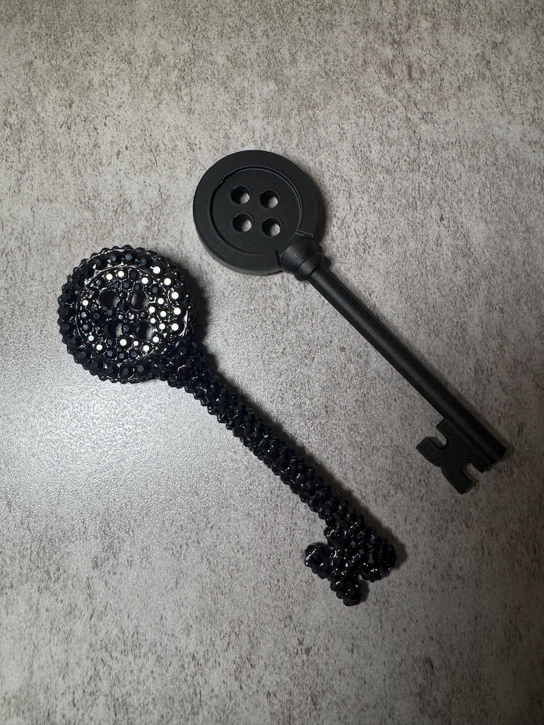 May include: Two decorative keys. One key has a button-shaped head with four holes and a long stem. The other key has a round head covered in black gems and a stem covered in black beads.