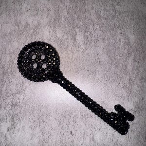 May include: A black decorative key with a detailed design. The key is covered in small, dark beads, giving it a textured appearance. The key's head has a circular shape with several holes.