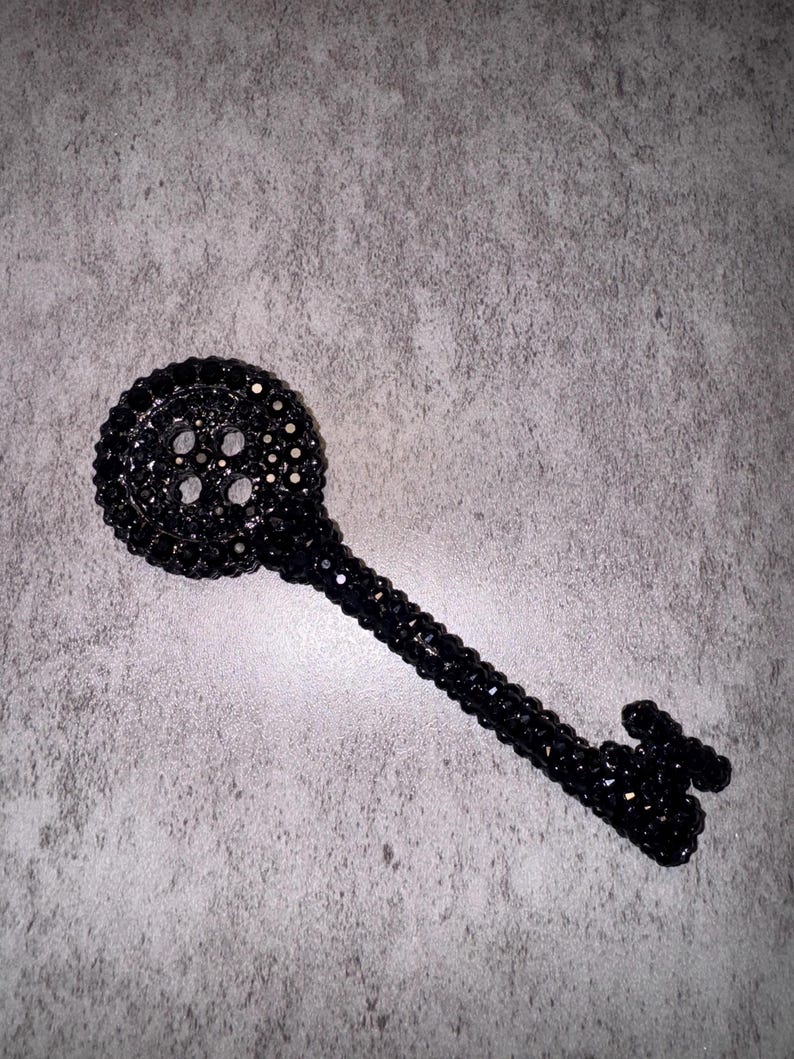 May include: A black decorative key with a detailed design. The key is covered in small, dark beads, giving it a textured appearance. The key's head has a circular shape with several holes.