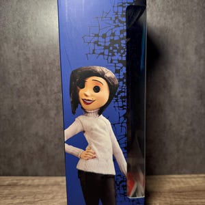May include: Ages 14 & Up. Boxed doll featuring a character with black hair, a white turtleneck sweater, and black pants. The box has a blue background with a graphic design and a clear plastic window. The doll is a collectible item.