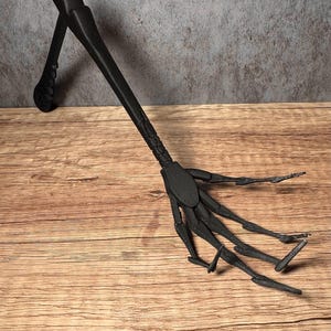 May include: A black, articulated wooden hand prop, with a long arm and a detailed, textured design. The hand is posed with fingers extended, and the arm is angled. The prop is set against a wooden surface.