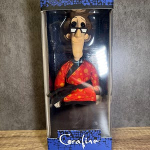 May include: A doll of a character from the movie "Coraline" in a clear plastic box. The doll wears glasses, a red jacket with orange polka dots, and black pants. The box has the movie title "Coraline" in blue.