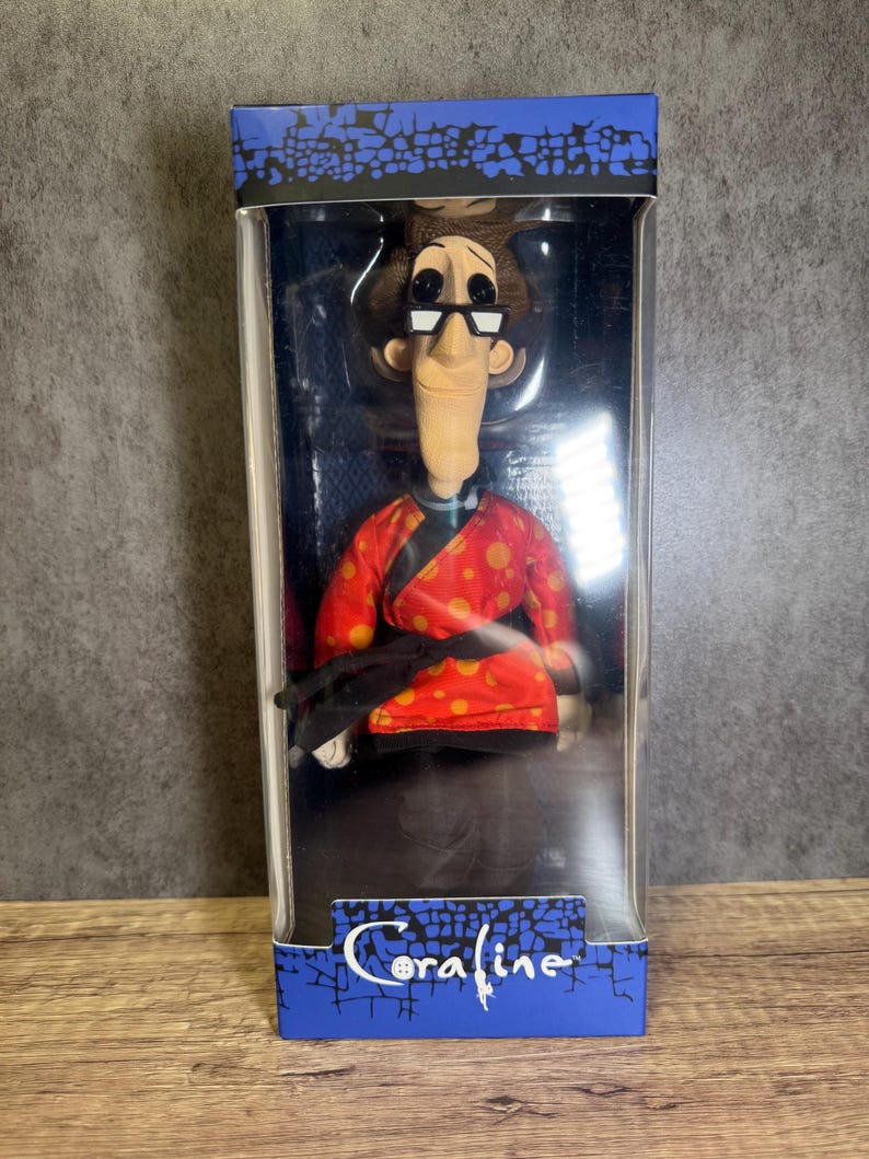May include: A doll of a character from the movie "Coraline" in a clear plastic box. The doll wears glasses, a red jacket with orange polka dots, and black pants. The box has the movie title "Coraline" in blue.