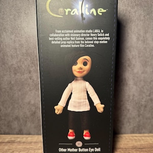 May include: A box featuring the Coraline movie doll, Other Mother Button Eye Doll. The doll is wearing a white sweater, black pants, and red shoes. The box has the Coraline logo and text from the movie.
