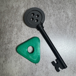 May include: A black key with a circular top featuring four holes, and a green triangular object with a central hole. The key has a long, straight handle and a detailed key bit. The objects are set against a gray textured background.