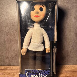 May include: A doll of a character from the movie "Coraline" in a clear plastic box. The doll has black button eyes, red lips, and black hair. She wears a white turtleneck sweater and brown pants. A black key is in front of the doll.