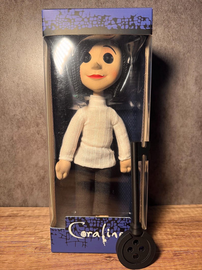 May include: A doll of a character from the movie "Coraline" in a clear plastic box. The doll has black button eyes, red lips, and black hair. She wears a white turtleneck sweater and brown pants. A black key is in front of the doll.