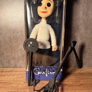 May include: A doll of a character from the movie "Coraline" in a clear plastic box. The doll has black hair, a white sweater, and black button eyes. A black key and a black hand-like object are in front of the box.