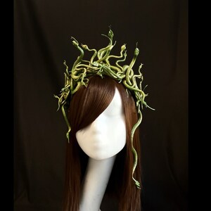 Puede incluir: Una diadema inspirada en Medusa con una corona de serpientes verdes. Las serpientes est&aacute;n detalladas y parecen enrolladas. La diadema se exhibe en una cabeza de maniqu&iacute; blanca con cabello casta&ntilde;o, sobre un fondo negro.