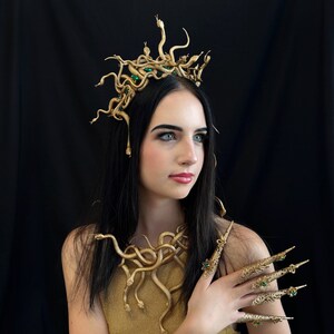 Puede incluir: Un conjunto de disfraz dorado con tem&aacute;tica de Medusa, que incluye una corona, un collar y garras para los dedos. La corona y el collar presentan dise&ntilde;os de serpientes con detalles verdes. Las garras tambi&eacute;n son doradas con gemas verdes.