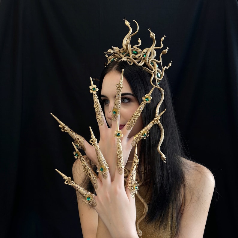 Puede incluir: Un conjunto de corona y u&ntilde;as doradas con el tema de Medusa. La corona presenta m&uacute;ltiples figuras de serpientes y piedras preciosas verdes. El conjunto de u&ntilde;as tiene intrincados dise&ntilde;os florales y piedras preciosas verdes. El fondo es negro.