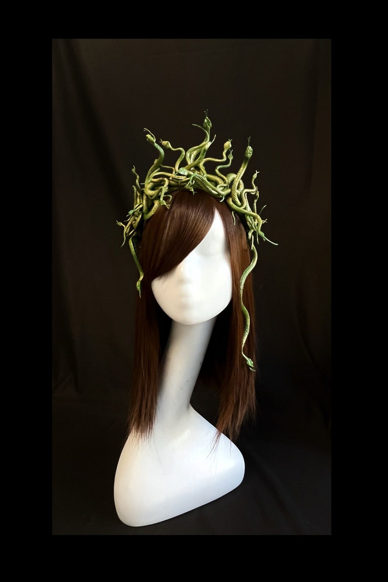 Puede incluir: Una diadema inspirada en Medusa con una corona de serpientes verdes. Las serpientes est&aacute;n detalladas y parecen enrolladas. La diadema se exhibe en una cabeza de maniqu&iacute; blanca con cabello casta&ntilde;o, sobre un fondo negro.