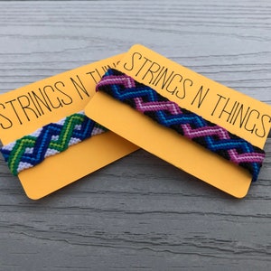 May include: Two friendship bracelets with chevron patterns on yellow cards with the text "Strings N Things". The bracelets are made with blue, green, pink, white, and black embroidery floss.