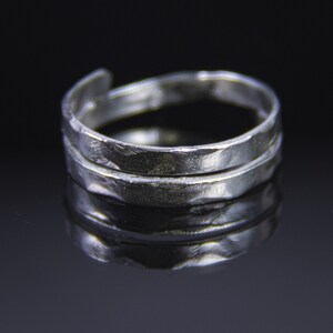 May include: A silver-colored ring with a textured, hammered appearance. The ring features a spiral design with two main bands and an adjustable opening. The ring is displayed against a dark background, highlighting its reflective surface.