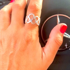 Puede incluir: Un anillo plateado con un diseño abstracto abierto con bucles interconectados. El anillo está en un dedo con esmalte de uñas rojo, apoyado en un volante negro con el logotipo de Mercedes-Benz.