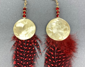 May include: A pair of gold-colored earrings featuring a hammered disc design, red crystal beads, and dark red and black spotted feathers. The earrings have gold-colored fish hook ear wires.