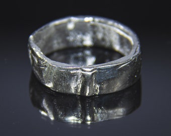 Könnte beinhalten: Ein silberner Ring mit einer strukturierten, gehämmerten Oberfläche. Das Band ist dick und hat eine leicht unregelmäßige Form, die das Licht reflektiert. Der Ring wird vor einem dunklen Hintergrund gezeigt, um seinen metallischen Glanz hervorzuheben.