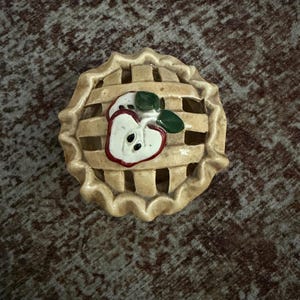 May include: A round ceramic pie-shaped container with a lattice-style lid. The lid features a sculpted, hand-painted apple slice with a red outline, white flesh, and green leaves. The crust is a light tan color with a scalloped, crimped edge design.