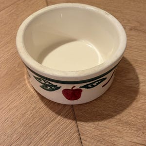 May include: A round, off-white ceramic bowl featuring a painted decorative border around the exterior. The design includes red apples and green leaves set against a thin dark green horizontal line, with a smooth, cream-colored interior finish.