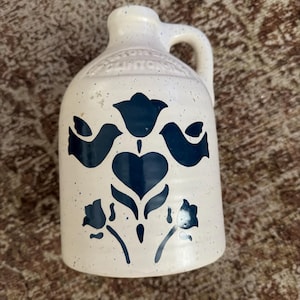 May include: A cream-colored ceramic jug with a speckled finish and a single handle. The front features a dark blue graphic design of two birds, a central heart, and floral motifs. Embossed text near the neck reads Clinton Iowa.