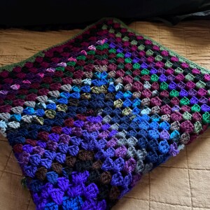 May include: A square crochet blanket featuring a multi-colored granny square pattern. The design incorporates a vibrant spectrum of yarn colors including deep blues, purples, greens, and black, arranged in a dense, textured, repeating cluster stitch.