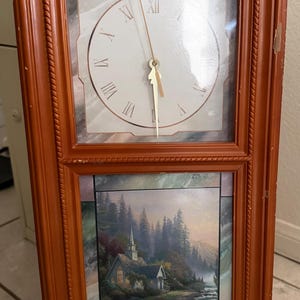 May include: A rectangular, wooden-framed clock with a dual-panel design. The top panel features a clock face with Roman numerals. The bottom panel displays a landscape painting of a church and river, with the word "Autumn" at the bottom.