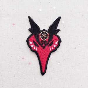 Puede incluir: Parche bordado con un diseño estilizado de criatura cornuda roja y negra. El parche tiene una cara roja con detalles blancos y cuernos negros. Una forma geométrica dorada está bordada en el centro superior. Para ropa o accesorios.