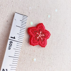 Puede incluir: Un parche de flor bordada roja con centro blanco y beige, de aproximadamente 2,5 cm de diámetro. El parche está colocado junto a una regla marcada en pulgadas, sobre un fondo de tela beige.