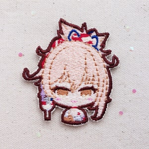 May include: Embroidered patch featuring a cartoon character with brown hair, wearing a red and blue headband. The character has brown eyes and a pink blush. The patch is on a white fabric background.