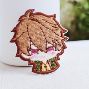 May include: Embroidered patch of a cartoon character with brown hair, pink eyes, and a green and yellow shirt.